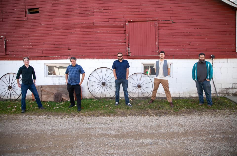 The band poses in a socially distanced manner in front of a barn