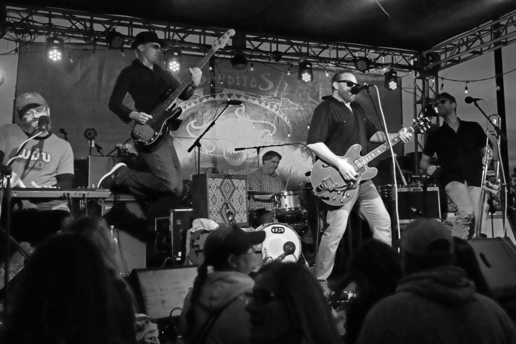 Black and white photo of keyboardist, bassist leaping, drummer, guitarist, and trombonist on an outdoor stage