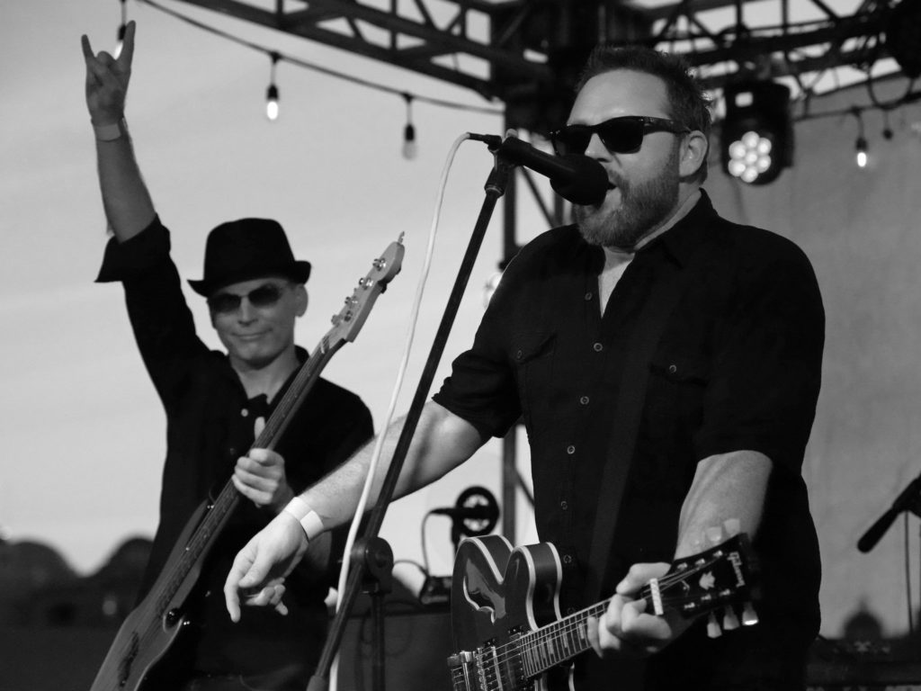 Black and white photo of bass player making the "horns up" sign behind singer/guitarist
