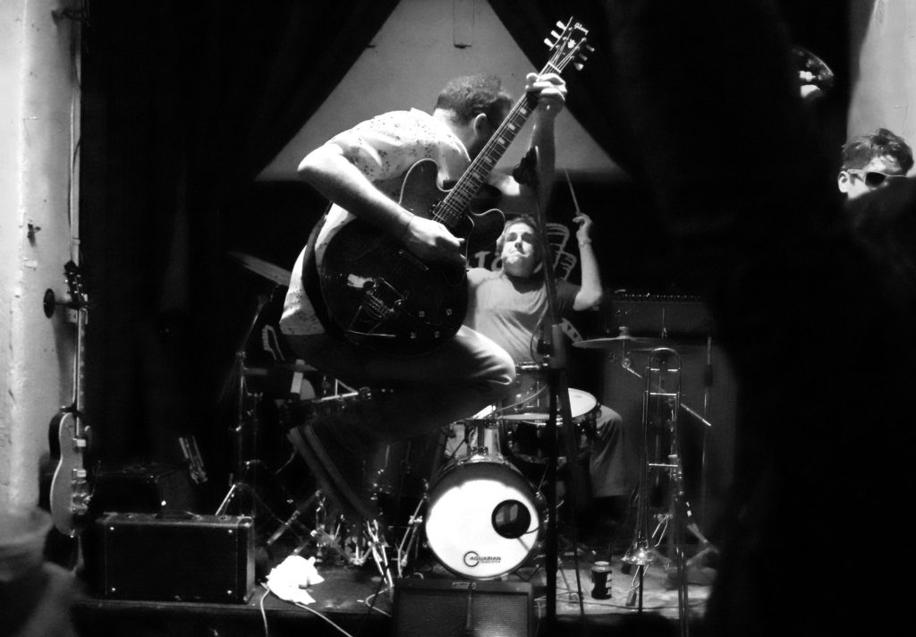 Black and white photo of guitarist leaping with drummer in background